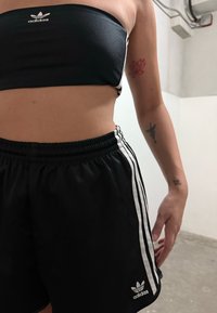 Oseba v črnem športnem modrčku in kratkih hlačah Adidas, ki kaže tetovaže na roki, stoji v industrijskem notranjem prostoru z betonskim tlakom.