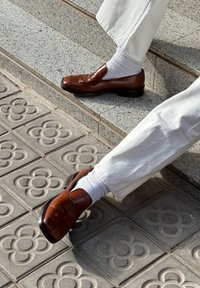 Rjavi usnjeni loafers z vtkanim vzorcem, kombinirani z belimi gležnjarskimi nogavicami, na stopnišču zz teksturiranimi sivimi ploščicami.