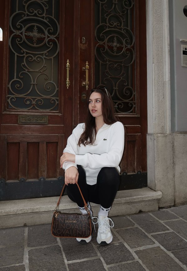 Mlada ženska z rjavimi lasmi, oblečena v belo majico z dolgimi rokavi, črne pajkice in bele superge, počepa na kamnitih stopnicah in drži rjavo torbico z vzorcem.