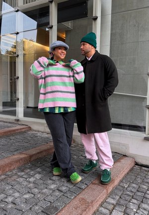 Dos personas están de pie en escalones de adoquines frente a un edificio; una lleva un suéter a rayas rosa y verde y un sombrero gris, la otra un abrigo negro y un gorro verde.