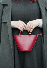 Femme portant une robe et un manteau gris foncé, tenant un sac à main trapézoïdal rouge  à deux mains, orné de bagues en or et d'un bracelet.