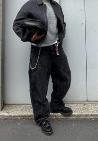 Pantaloni ampi in corduroy neri con dettaglio in catena argentata, maglione grigio a maglia, giacca bomber scura e scarpe in pelle nere.