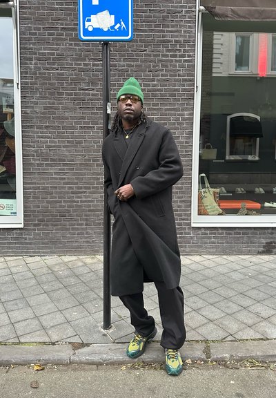 Abrigo negro de doble botonadura sobre un conjunto negro, con un gorro verde y zapatillas con un patrón verde y amarillo. De pie al lado de un cartel.
