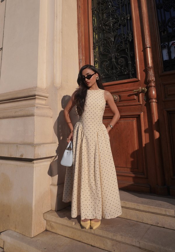 Vrouw draagt een crèmekleurige mouwloze polkadotjurk, gele hakken, zwarte zonnebril en houdt een witte handtas vast, staand bij een houten deur.