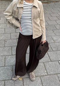Giacca in suede sintetico beige con frange, pantaloni neri a gamba larga, camicia a righe, scarpe fantasia e borsa intrecciata marrone. La superficie del terreno è in pietra.