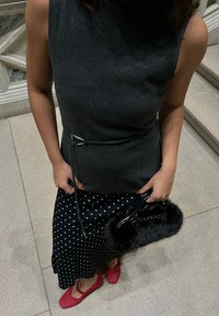Femme portant un haut sans manches gris foncé avec ceinture, jupe noire à pois blancs, chaussures rouges, tenant un petit sac à main noir poilu à l'intérieur.