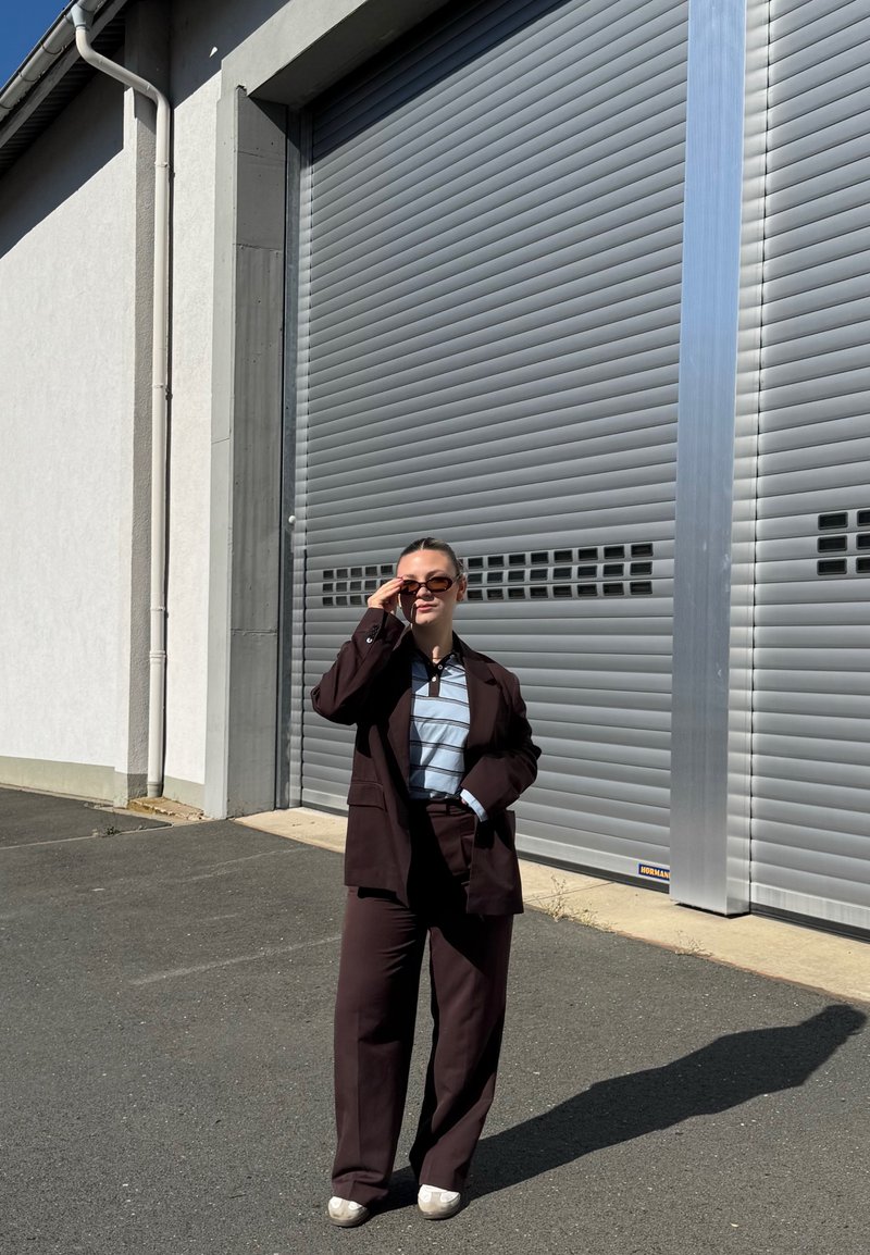 Femme portant un costume sombre et une chemise rayée ajuste ses lunettes de soleil, debout devant une grande porte industrielle métallique fermée à volet, en plein jour.