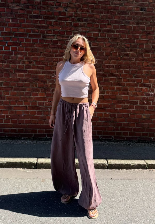 Jonge vrouw met zonnebril, een wit mouwloos crop top en losse paarse broek, staand op het trottoir tegen een rode bakstenen muur in het zonlicht.