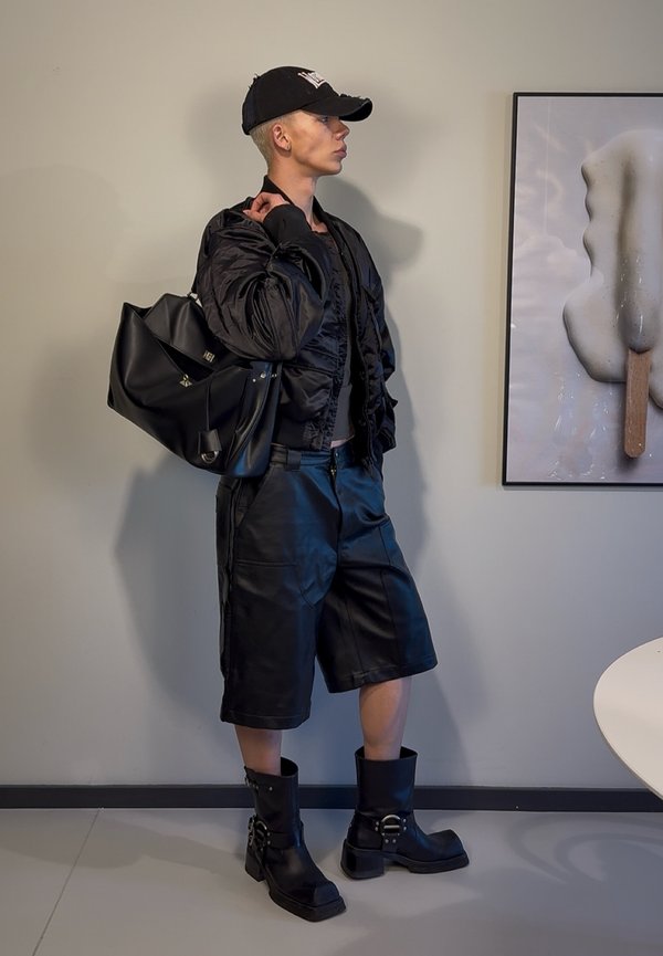 Zwarte bomberjack over een aansluitend zwart topje, gecombineerd met zwarte leren shorts en kniehoge laarzen. Het model houdt een matte zwarte tas vast.