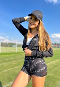 Crna sportska jakna s mrežastim detaljima, bijela majica bez rukava i crne šortsevi s uzorkom Nike logoa. Vanjska okolina s plavim nebom.