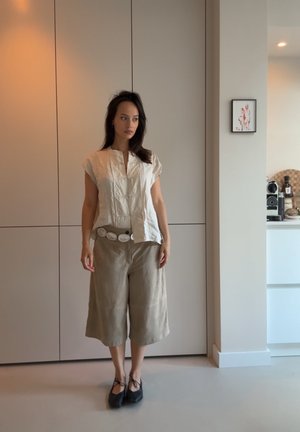 Blusa de algodón color hueso con mangas cortas y escote en V, combinada con culottes beige y un cinturón ancho con acentos circulares. Zapatos negros.