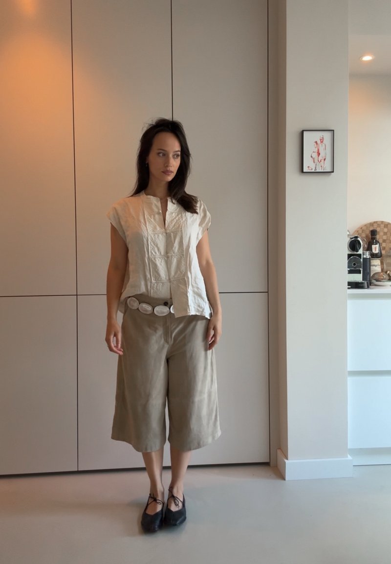 Blouse en coton blanc cassé à manches courtes et décolleté en V, associée à des culottes beiges et une large ceinture avec des accents circulaires. Chaussures noires.