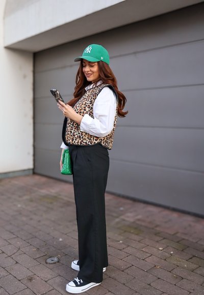 Chaleco de estampado de leopardo sobre una camisa blanca, pantalones de pierna ancha negros, gorra verde y bolso acolchado verde. Lleva puesta unas zapatillas negras y blancas. Usando un teléfono.