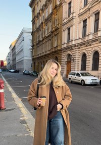 Long manteau en laine brune sur un haut foncé, jean bleu, tenant une tasse de café. Cadre urbain avec bâtiments historiques et voitures stationnées.