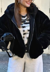 Chaqueta de piel sintética negra con cierre de cremallera, textura suave y capucha. Lleva puesta sobre una camiseta gráfica de rayas y combinada con pantalones de chándal grises.