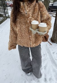 Personne portant une veste en fausse fourrure marron à taches et un pantalon gris, tenant deux gobelets de café à emporter dans un cadre extérieur enneigé.