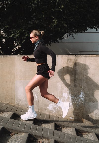 Mujer vestida con ropa deportiva negra y zapatillas blancas corriendo por unas escaleras exteriores de hormigón junto a una pared.