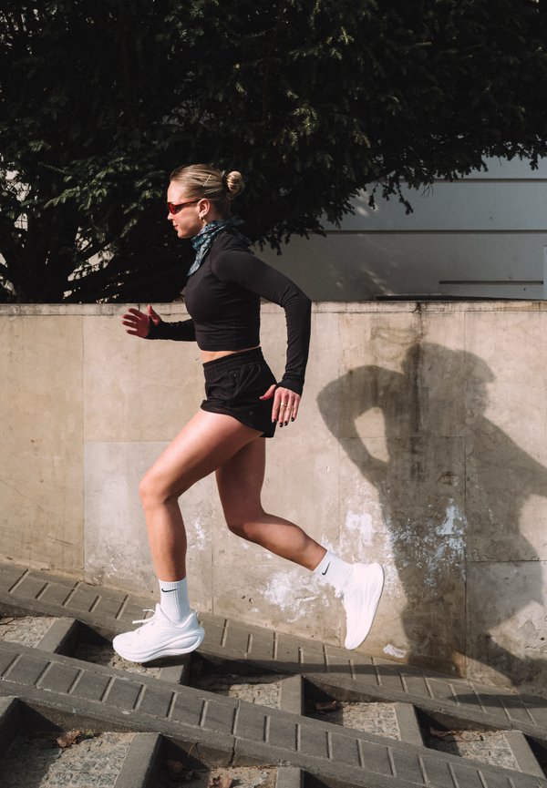 Kobieta ubrana w czarne sportowe ubrania i białe trampki biegnie po zewnętrznych betonowych schodach obok ściany.