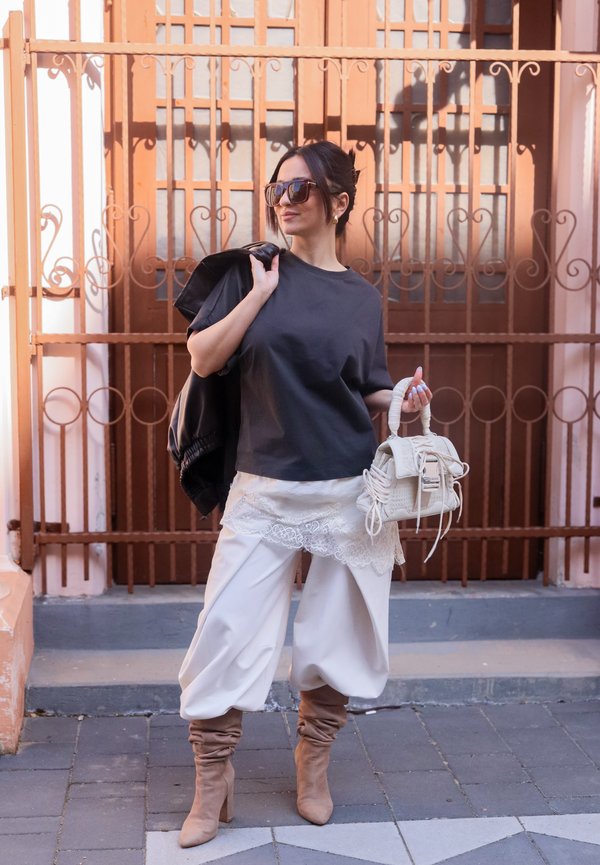 Femeie cu ochelari de soare supradimensionați, cămașă neagră, pantaloni albi cu dantelă, cizme bej, ținând o jachetă neagră și o geantă albă pe o stradă din oraș.