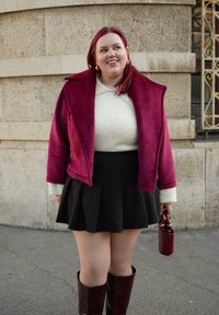 Jachetă din piele de căprioară burgundy, pulover tricotat crem, fustă plisată neagră și cizme lungi până la genunchi din piele maro. Ține o geantă mică burgundy.