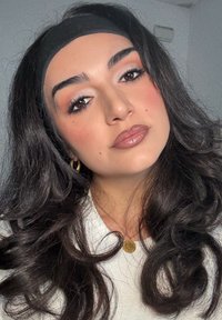 De longs cheveux noirs avec des boucles lâches, portant un serre-tête noir, un pull de couleur claire et un collier avec un pendentif circulaire en or. Maquillage subtil.