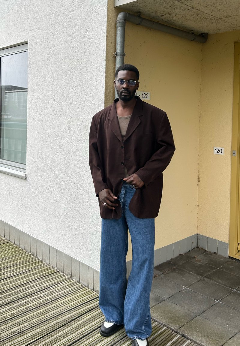 Brauner übergroßer Blazer, beigefarbenes Top, weite blaue Jeans und schwarz-weiße Schuhe. Steht auf einer strukturierten Fläche in der Nähe einer gelben Wand.
