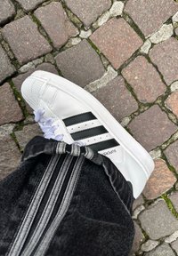 Baskets Adidas Superstar blanches avec des bandes noires sur un pavé en pierres, portées avec un pantalon noir avec des bandes grises.