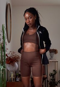 Brązowy sportowy crop top i szorty z wysokim stanem wykonane z gładkiego materiału. Noszone z czarną kurtką na zamek. Wewnętrzna przestrzeń w otoczeniu roślinności.