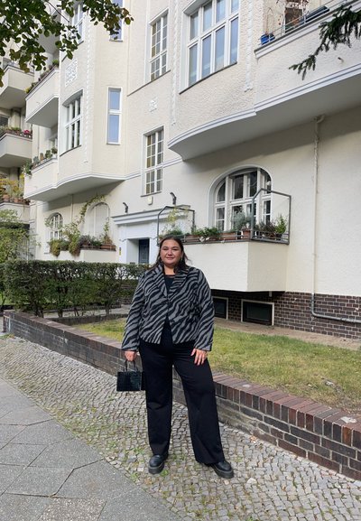 Chaqueta a rayas de cebra, camiseta negra, pantalones negros y bolso negro. De pie en un camino de adoquines con edificios residenciales al fondo.