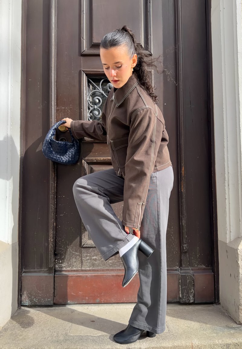 Femme en veste marron et pantalon gris ajustant une botte noire à talon tout en tenant un petit sac à main bleu, se tenant devant une porte en bois.