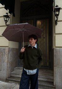 Homme aux cheveux bouclés debout sur des marches en pierre tenant un parapluie marron ouvert, portant un pull sombre par-dessus une chemise rayée et un pantalon bleu.