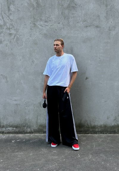 Camiseta de algodón blanca, pantalones de chándal negros holgados con franjas blancas, zapatillas rojas, sosteniendo auriculares, contra una pared de concreto gris.