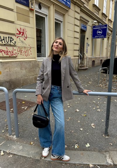 Blazer de cuadros grises de doble botonadura sobre un suéter negro de cuello alto, combinado con jeans azules de pierna ancha y zapatillas blancas. Sosteniendo un bolso negro.