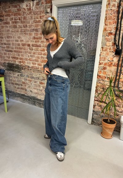 Cardigan de punto gris sobre una camiseta sin mangas blanca, combinado con jeans de denim azul holgados. El entorno presenta una pared de ladrillo y una planta en maceta cercana.