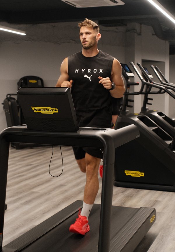 Homme courant sur un tapis de course Technogym en salle de sport, portant un débardeur noir et des chaussures de course rouges.