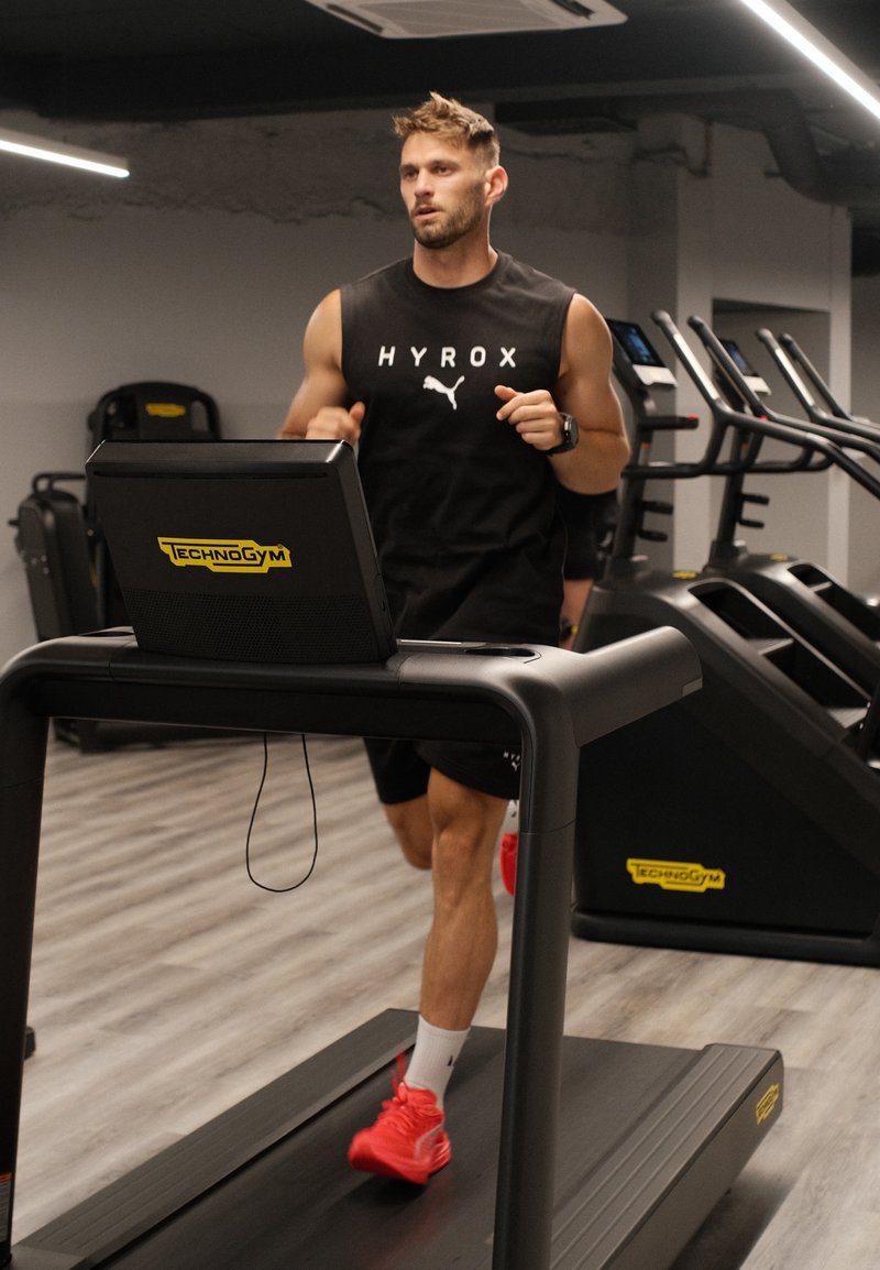 Homme courant sur un tapis roulant Technogym dans une salle de sport, portant un débardeur noir et des chaussures de course rouges.