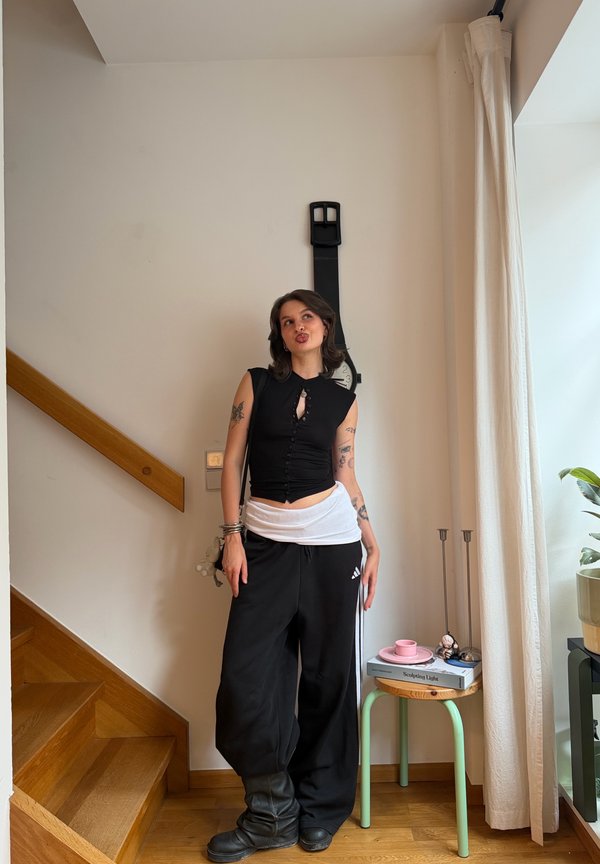 Femme avec des tatouages portant un top noir sans manches et un pantalon large noir, debout à l'intérieur près des escaliers et d'une petite table avec des livres et une plante.