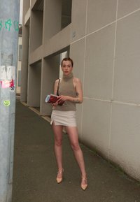 Neule, vaaleanruskea hihaton paita, yhdessä kermaisen minihameen kanssa, jossa on sivuhalkio. Pitelee kirjaa ja seisoo asfaltilla.