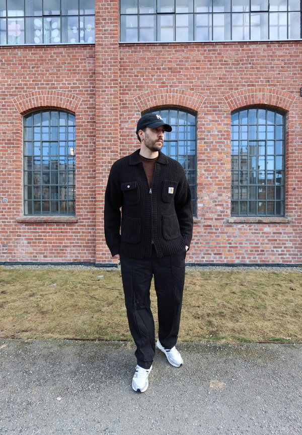 Homme portant un pull noir, un pantalon, une casquette de baseball et des baskets blanches, debout sur un chemin devant un bâtiment en brique avec de grandes fenêtres.