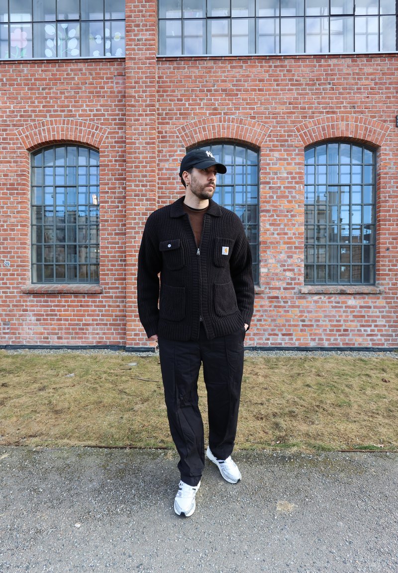 Homme portant un pull noir, un pantalon, une casquette de baseball et des baskets blanches, debout sur un chemin devant un bâtiment en briques avec de grandes fenêtres.