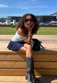 Γκρι μπότες μέχρι το γόνατο, ανοιχτό μπλε τοπ, και ναυτικό φούστα. Σγουρά μαλλιά, μικρές μαύρες ηλιαχτίδες, καθιστή σε ξύλινη πλατφόρμα. Μαύρη τσάντα κοντά.