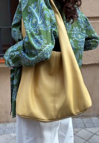 Saco hobo amarelo em pele com uma forma descontraída, textura suave e uma costura central, usado casualmente sobre uma blusa verde com padrão paisley.