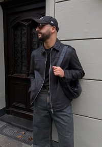 Černá kožená bunda se zipem, kombinovaná s černou košilí a šedými džíny. Doplněná černou taškou a čepicí. Dřevěné dveře v pozadí.