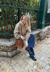 Beige trenčkot so širokými rukávmi, modré džínsy, čierne topánky a hnedá kožená taška. Leží na kamennej lavičke so jesennými listami.