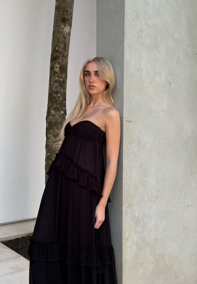Vestido maxi negro sin tirantes con volantes, diseño en capas y tela ligera. La modelo está de pie junto a un muro de piedra, con un árbol esbelto cerca.