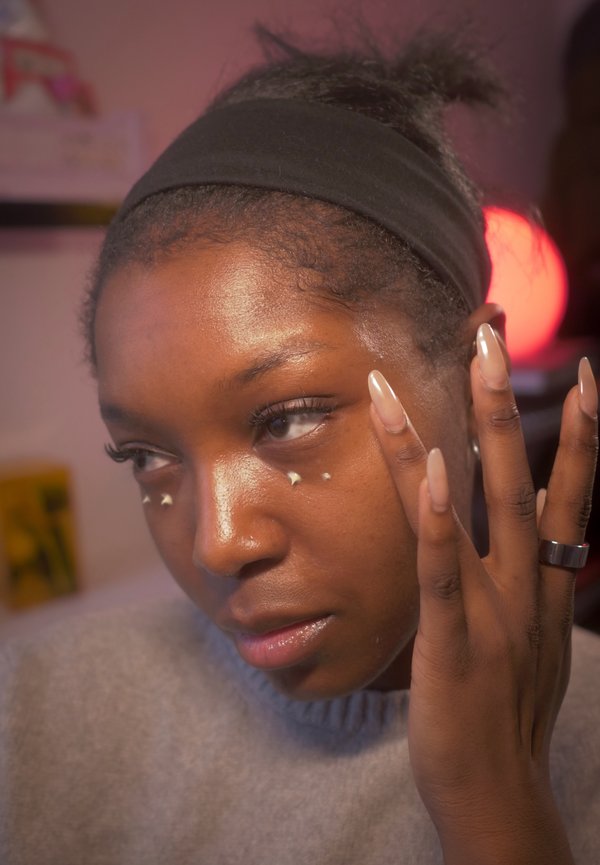 Model draagt een grijze trui en brengt huidverzorging aan. Nagels zijn lang en nude van kleur. Een zwart haarband houdt het haar uit het gezicht. Accentsterren bij de ogen.