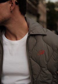 Veste grise matelassée avec une fermeture éclair et un logo rouge brodé, portée par-dessus un t-shirt blanc. Elle présente un col décontracté et une texture douce.