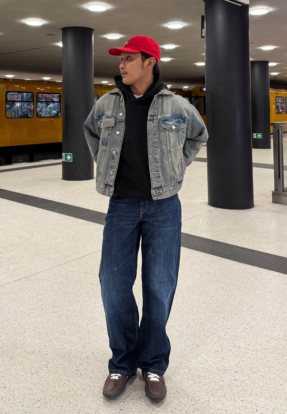 Veste en jean portée sur un sweat à capuche noir, jean bleu ample, et chaussures marron à lacets blancs. Casquette rouge portée à l'envers. Décor intérieur avec un fond de train jaune.