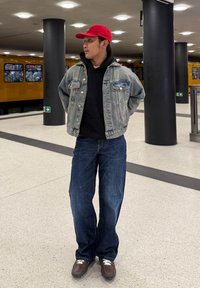 Veste en jean portée sur un sweat à capuche noir, jean bleu ample, et chaussures marron à lacets blancs. Casquette rouge portée à l'envers. Décor intérieur avec un fond de train jaune.