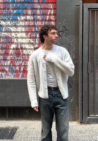 Λευκό, αφράτο κάρντιγκαν με σχέδιο μπροστά με κουμπιά πάνω από ένα ανοιχτό γκρι T-shirt, συνδυασμένο με σκούρα τζιν. Στηρίζεται σε έναν πολύχρωμο τοίχο.